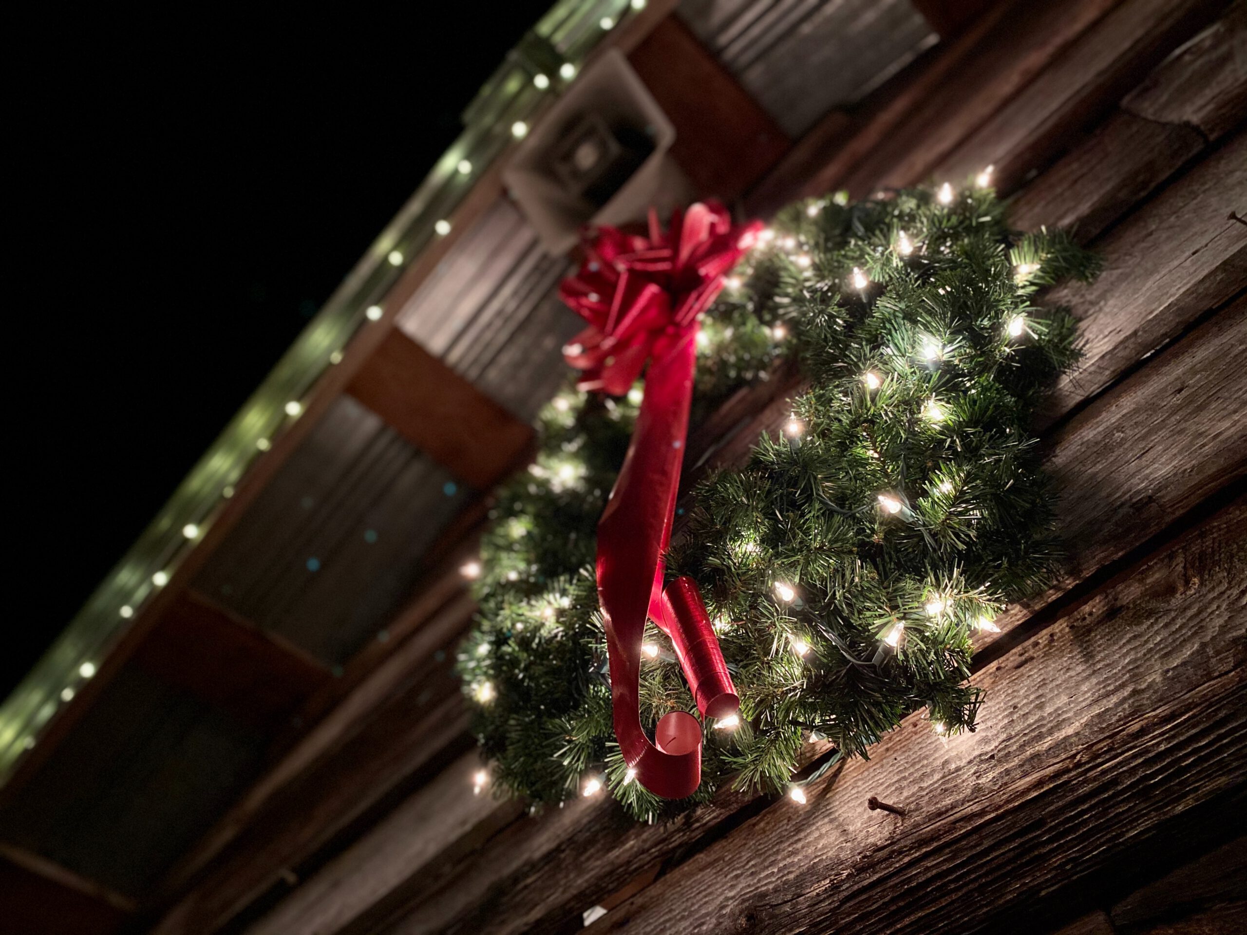 Zielony wieniec świąteczny ze świecącymi światełkami i czerwoną wstążką wiszący na drewnianej ścianie
